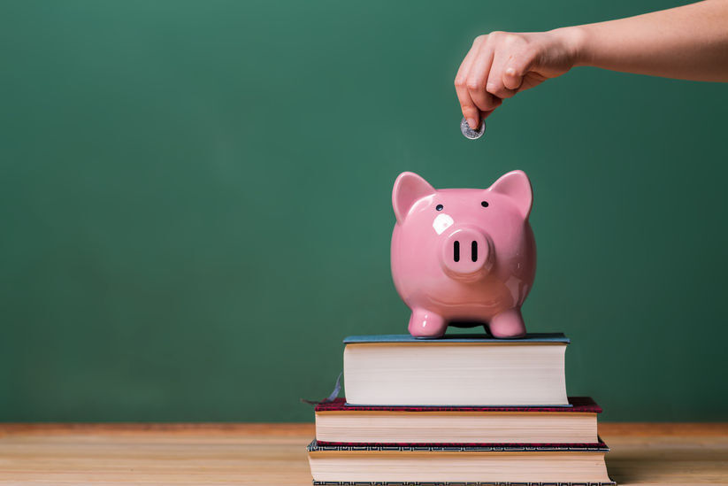 37509139 - person depositing money in a pink piggy bank on top of books with chalkboard in the background as concept image of the costs of education
